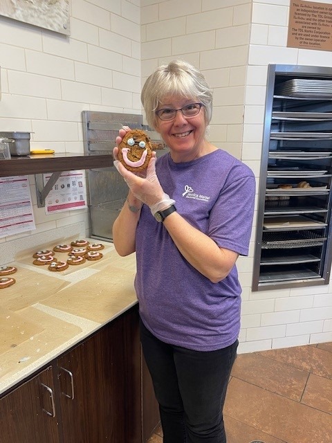 A Tim Hortons team member holding a Smile Cookie beside a tray of freshly iced cookies.